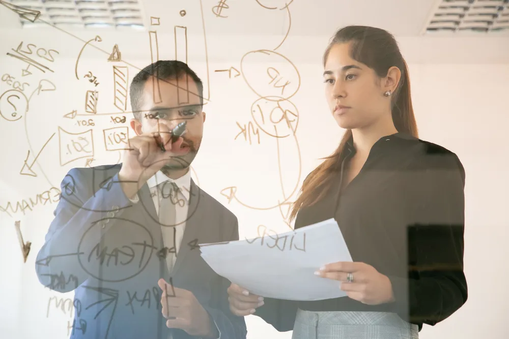 content-african-american-manager-writing-chart-glass-board-professional-young-pretty-female-colleague-holding-document-looking-graph-conference-room-teamwork-marketing-concept_
